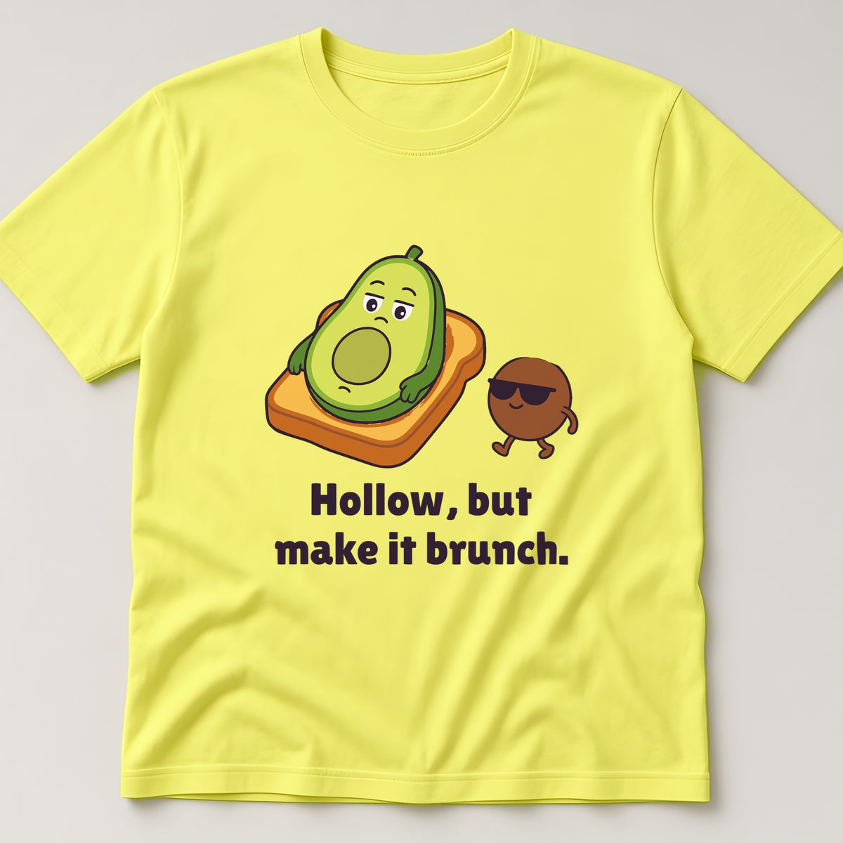 Cornsilk t-shirt, flat lay, printed with half an avocado on toast next to its pit in sunglasses and the words 'Hollow, but make it brunch.'