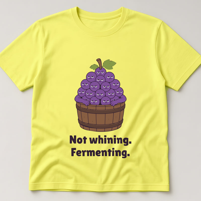 Cornsilk t-shirt, flat lay, printed with a bunch of grumpy grapes in a wooden barrel and the words 'Not whining. Fermenting.'