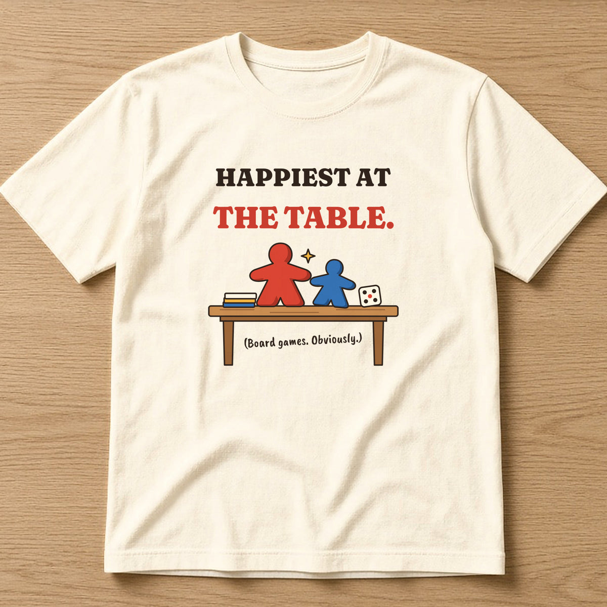 Natural t-shirt, flat lay, printed with the clean version: 'Happiest at the table. (Board games. Obviously.)'