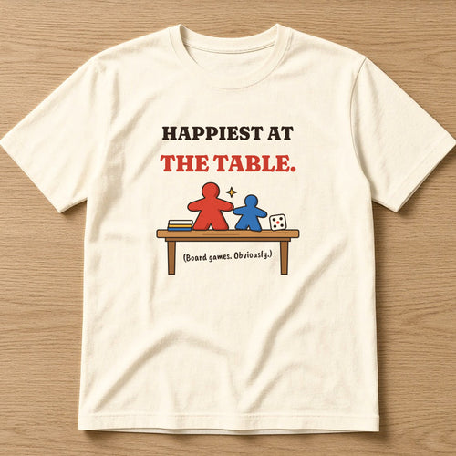 Natural t-shirt, flat lay, printed with the clean version: 'Happiest at the table. (Board games. Obviously.)'