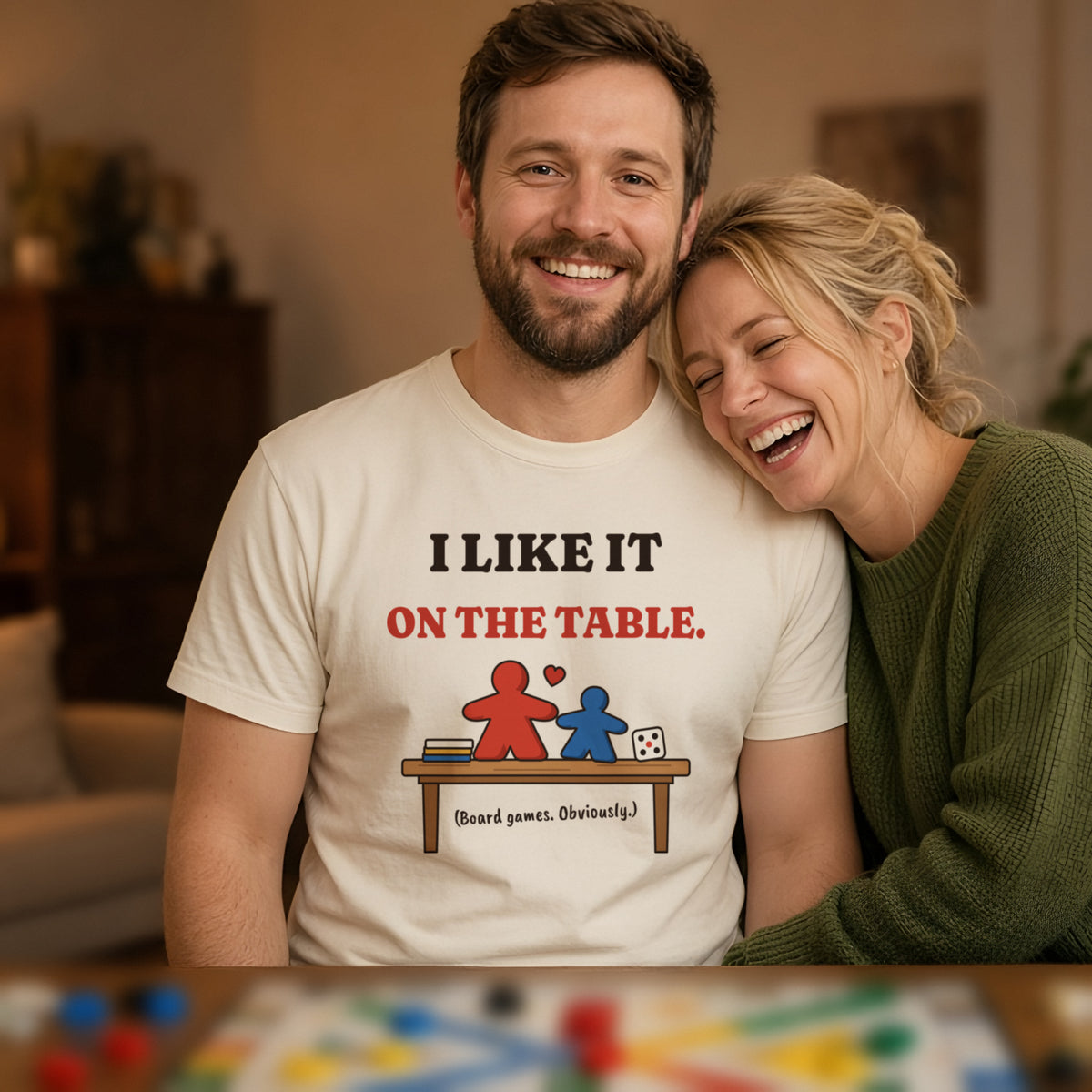 Man wearing a natural t-shirt printed with 'I like it on the table. (Board games. Obviously.)' (Spicy version), at a board game night. Model image is digitally generated. The print shown is the actual design; colours and fit may vary slightly, see size chart.
