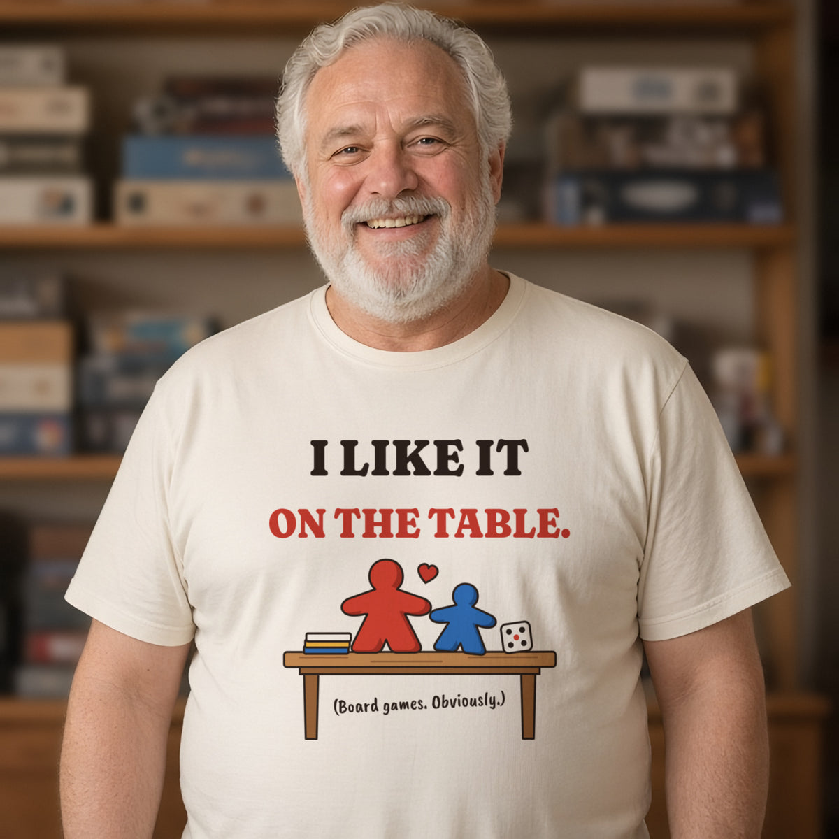 Man wearing a natural t-shirt printed with 'I like it on the table. (Board games. Obviously.)' (Spicy version), in a living room. Model image is digitally generated. The print shown is the actual design; colours and fit may vary slightly, see size chart.