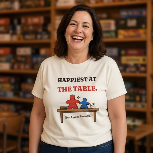 Woman wearing a natural t-shirt printed with 'Happiest at the table. (Board games. Obviously.)' (clean version), in a board game cafe. Model image is digitally generated. The print shown is the actual design; colours and fit may vary slightly, see size chart.