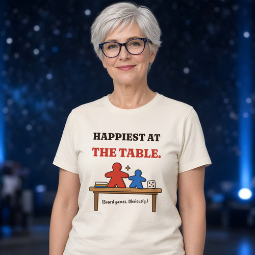 Woman wearing a natural t-shirt printed with 'Happiest at the table. (Board games. Obviously.)' (clean version), at a sci-fi convention. Model image is digitally generated. The print shown is the actual design; colours and fit may vary slightly, see size chart.