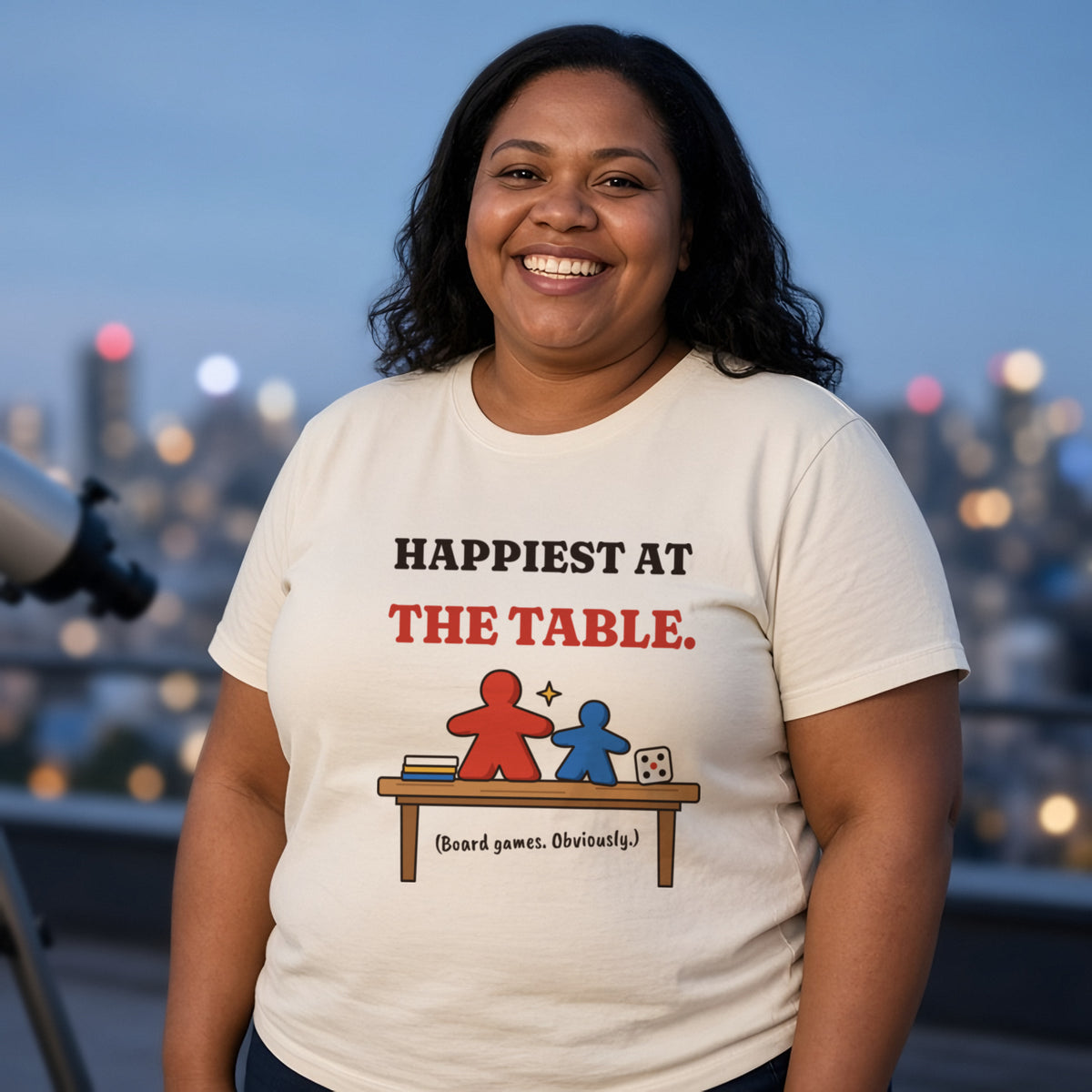 Woman wearing a natural t-shirt printed with 'Happiest at the table. (Board games. Obviously.)' (clean version), on a rooftop. Model image is digitally generated. The print shown is the actual design; colours and fit may vary slightly, see size chart.