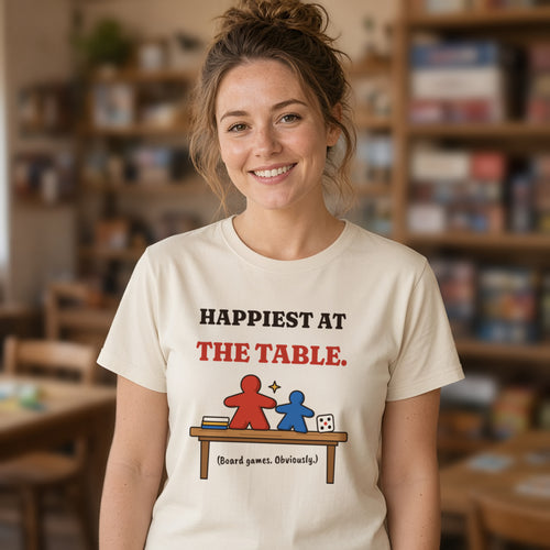 Woman in her twenties wearing a natural t-shirt printed with 'Happiest at the table. (Board games. Obviously.)' (clean version), at a games table. Colours and fit may vary slightly, see size chart.