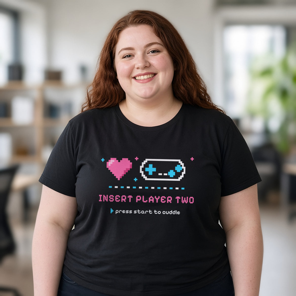 Woman wearing a black t-shirt printed with 'Insert Player Two / press start to cuddle' (Spicy version), in a co-working space. Model image is digitally generated. The print shown is the actual design; colours and fit may vary slightly, see size chart.