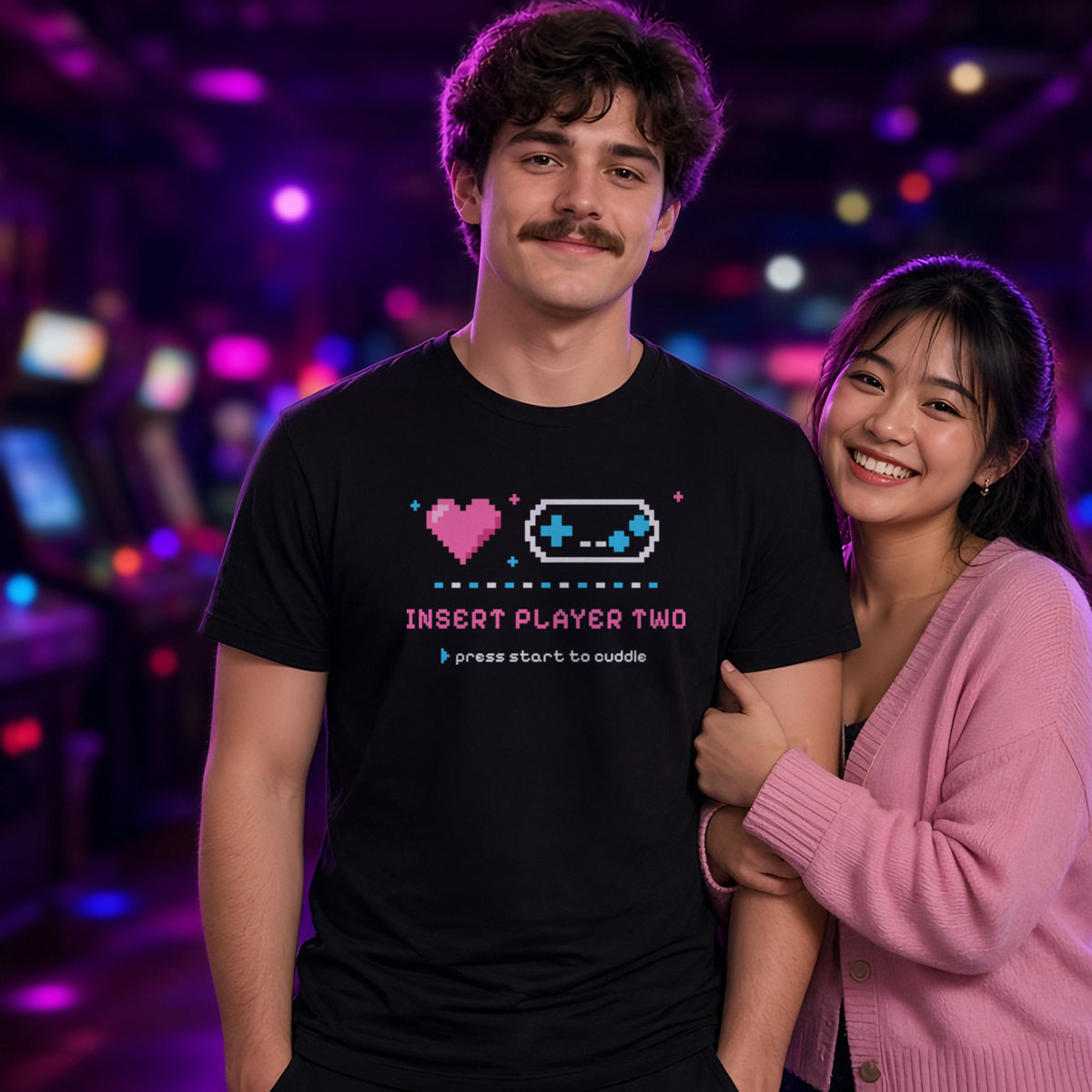 Man wearing a black t-shirt printed with 'Insert Player Two / press start to cuddle' (Spicy version), in an arcade bar. Model image is digitally generated. The print shown is the actual design; colours and fit may vary slightly, see size chart.