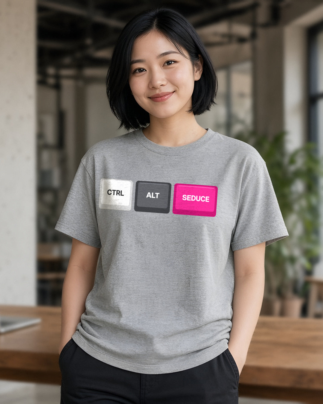 Woman wearing a sport grey t-shirt printed with Ctrl Alt Seduce keycaps (Spicy version), in a cafe (4:5 crop). Colours and fit may vary slightly, see size chart.